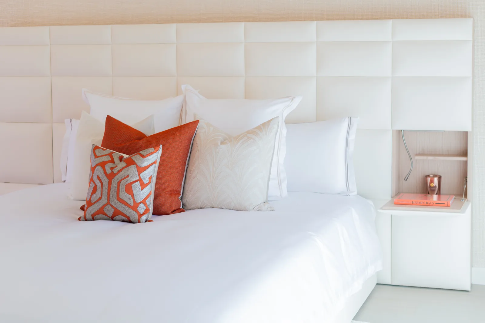 Cama con cabecero tapizado blanco, ropa de cama blanca y cojines en tonos naranja