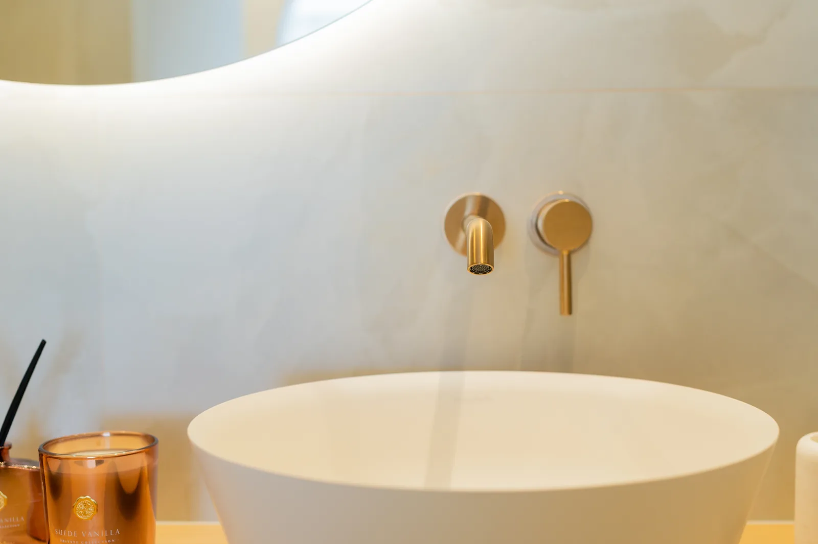 Lavabo redondo sobre encimera con grifería mural en oro cepillado y espejo curvo retroiluminado