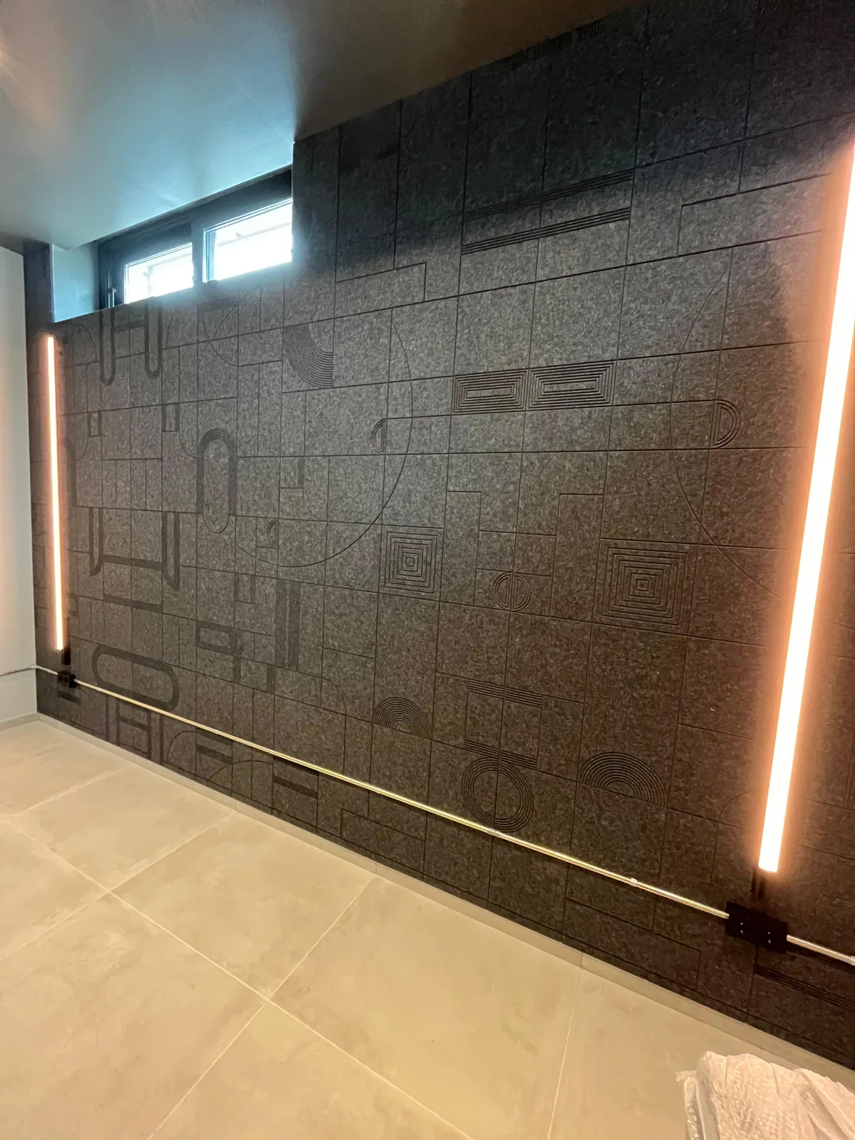 Pared completa de paneles acústicos de fieltro iluminada por dos tubos LED verticales y ventana alta