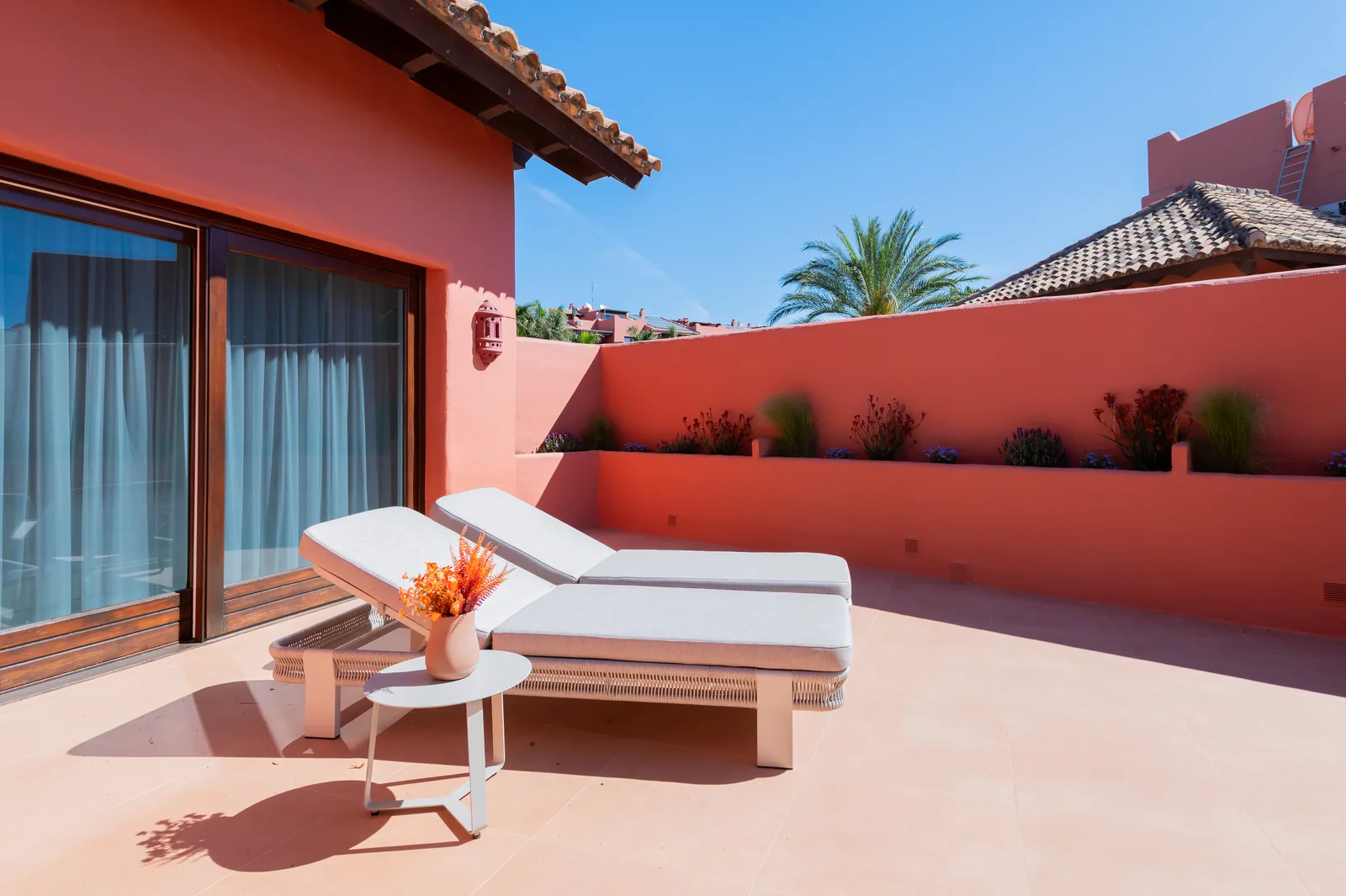 Terraza exterior con dos tumbonas, mesa auxiliar y jardineras corridas sobre muro coral